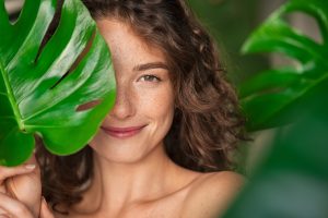 Close up face of beautiful young woman covering her face by green monstera leaf while looking at camera. Natural smiling girl with green palm leaf. Portrait of beauty woman with natural makeup and freckles on skin standing behind big green leaves.